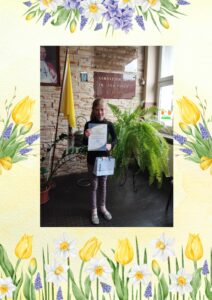 Uczennica w holu szkoły trzyma dyplom i nagrodę; w tle tablica szkoły, flaga i rośliny.