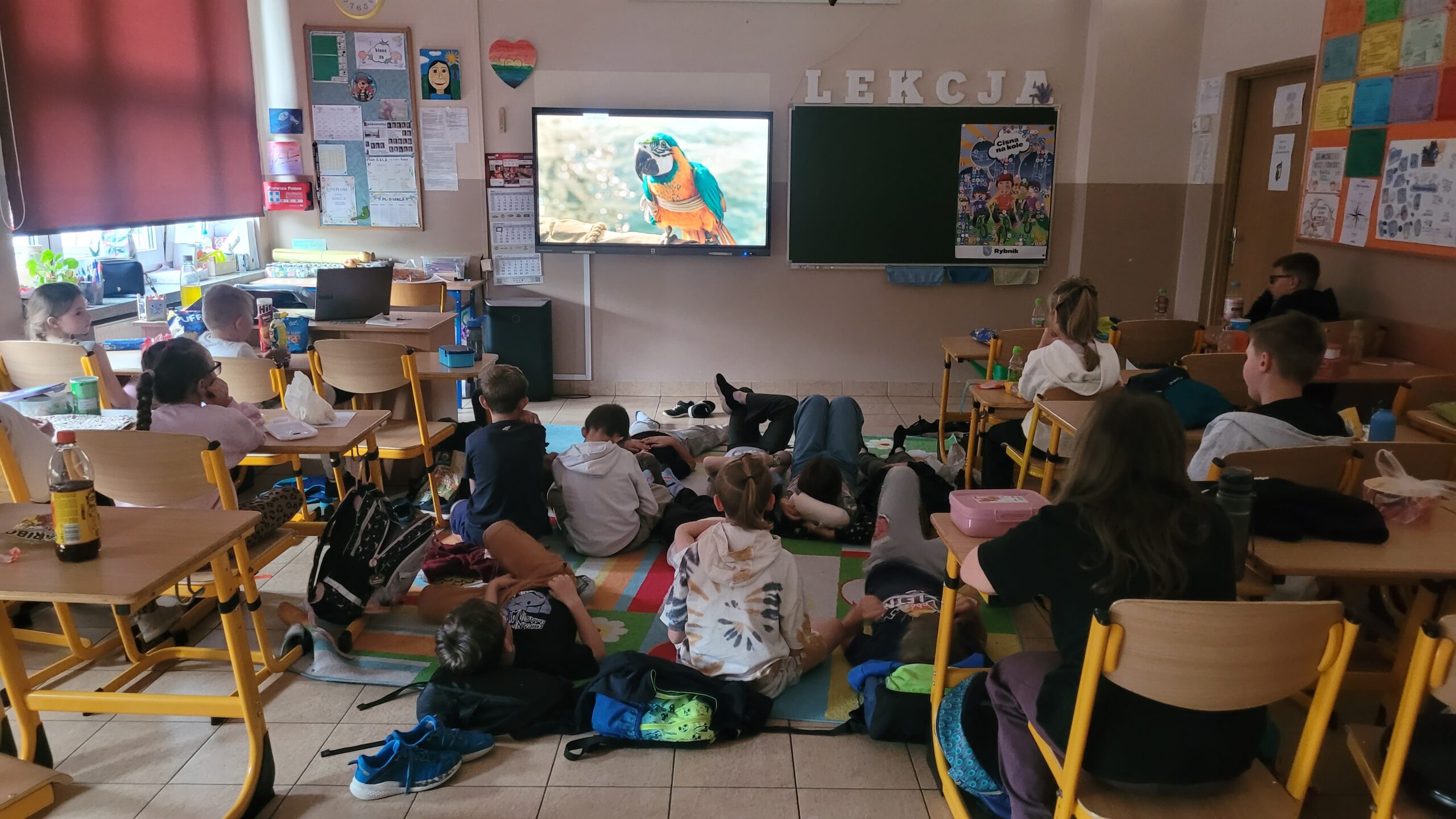 Dzieci uczestniczą w szkolnym dniu filmowym – siedzą przy ławkach i na dywanie, oglądając film wyświetlany na ekranie w klasie. W sali panuje swobodna atmosfera, uczniowie odpoczywają i wspólnie spędzają czas podczas projekcji.