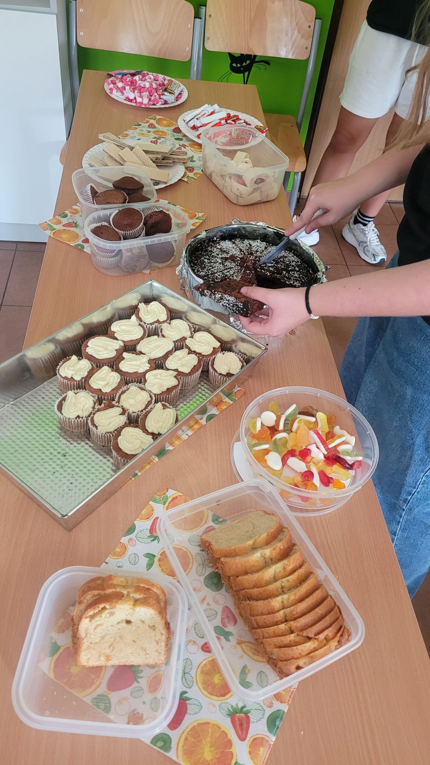 Zdjęcie przedstawia stół w sali szkolnej zastawiony domowymi wypiekami i słodyczami. Widać babeczki z kremem, kawałki ciasta czekoladowego, ciasto ucierane, wafle, cukierki i żelki. Przy stole stoją uczniowie, a jedna osoba nakłada kawałek ciasta.